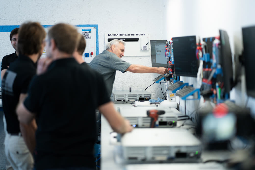 Ein Mitarbeiter zeigt Besuchern das KANBAN-Bestellboard in der Technik-Werkstatt