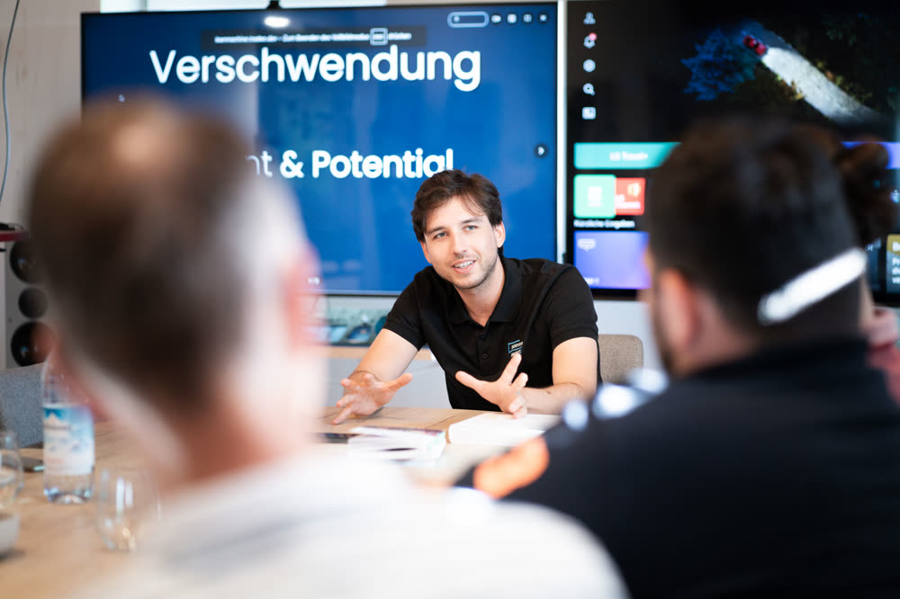 Ferdi präsentiert vor einem Screen mit der Slide „Verschwendung“ im Konferenzraum
